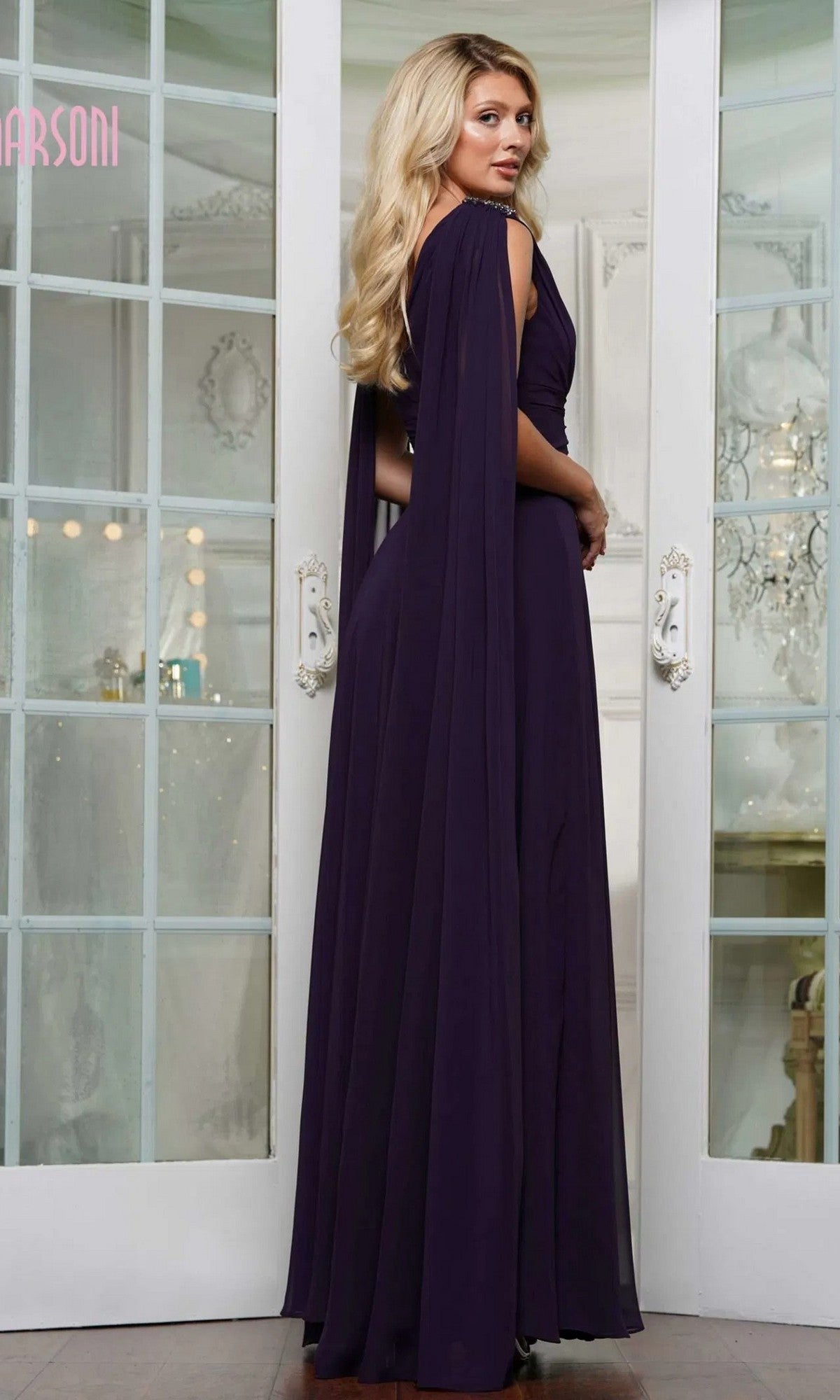 A woman with long blonde hair wears the Marsoni MV1349 floor-length deep purple formal dress with cape sleeves, standing in front of glass-paneled double doors.